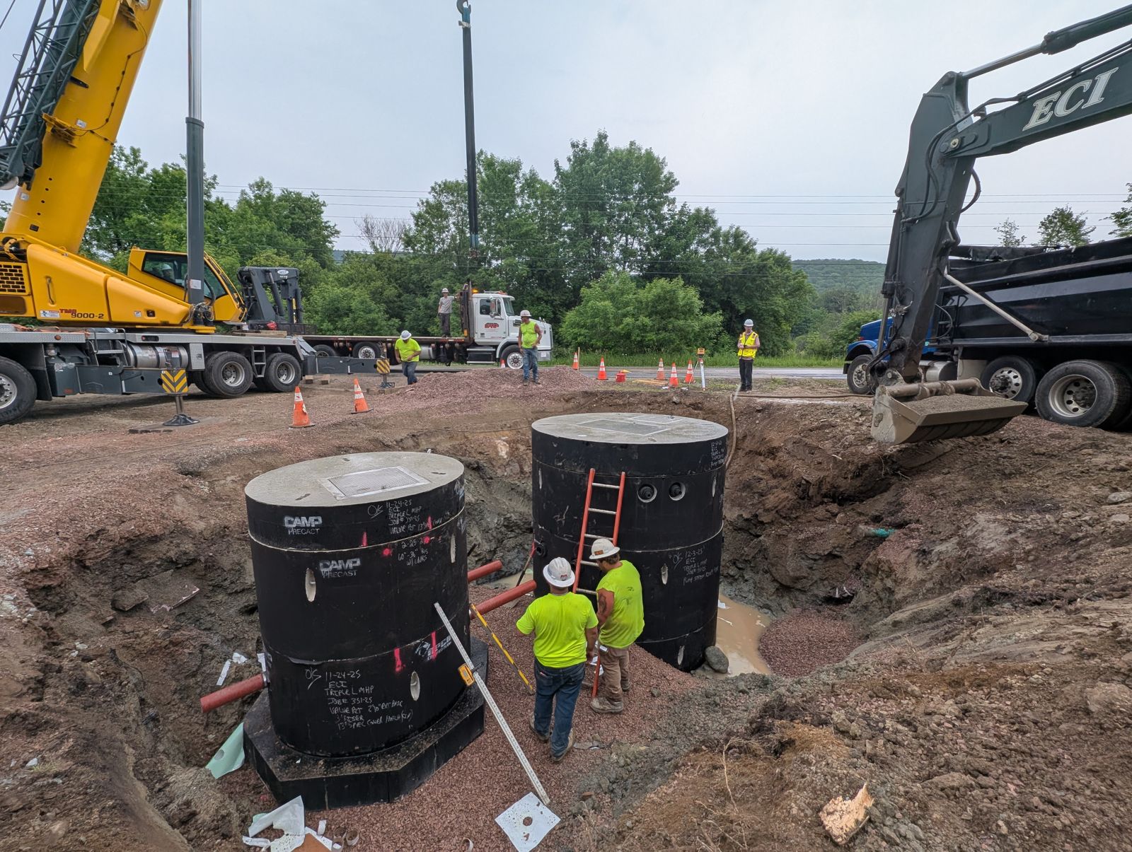Crane and excavator beside two large precast structures set in an excavation, crew working below